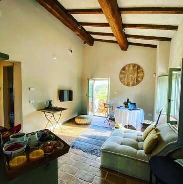 The image depicts a spacious room with a high ceiling, featuring a couch, table, and TV, with a doorway leading to a patio and a scenic view of the outdoors.