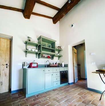 The image depicts a kitchen with light blue cabinets, a sink, and an oven, featuring wooden beams on the ceiling and a tiled floor, with a doorway leading to another room.
