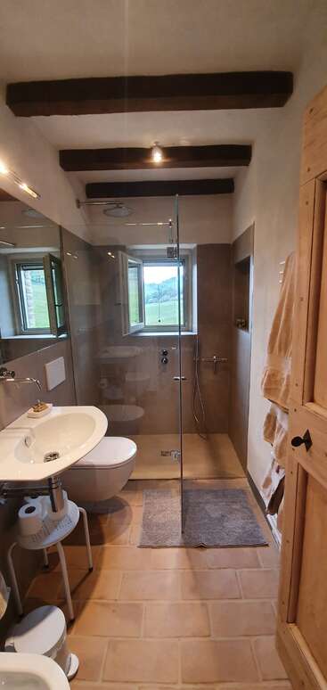 The image depicts a bathroom with a shower, sink, and toilet, featuring white walls and a tiled floor, with a large mirror above the sink and a window on the right side.