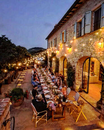 Les invités sont assis à une longue table à côté d'un bâtiment rustique en pierre et profitent d'une soirée chaleureuse.
