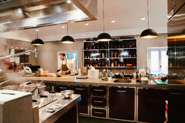 Un chef travaille dans une cuisine moderne et bien équipée, avec des étagères d'ingrédients, sous d'élégantes lampes suspendues, entouré de divers ustensiles, appareils et casseroles fumantes.