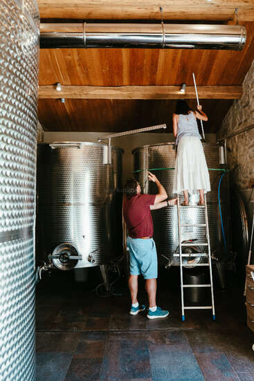 Une femme, debout sur une échelle, inspecte une grande cuve de fermentation en métal, tandis qu'un homme pointe du doigt et observe dans une salle de brassage ou de vinification rustique.