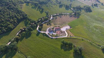Cette image aérienne montre une villa de campagne entourée de champs verts, d'arbres et d'une route sinueuse. On y voit des terres agricoles, des jardins paysagers et de vastes paysages naturels.