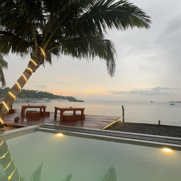 Une vue sereine au bord de la plage avec une piscine lumineuse, un palmier enveloppé de lumières, des sièges en bois sur la terrasse et les eaux calmes de l'océan au coucher du soleil. Une vue paisible et accueillante.
