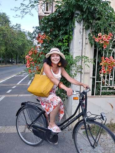 A woman in a hat and floral skirt sits on a black bicycle, with a yellow bag slung over her shoulder, in front of a white wall and a road.