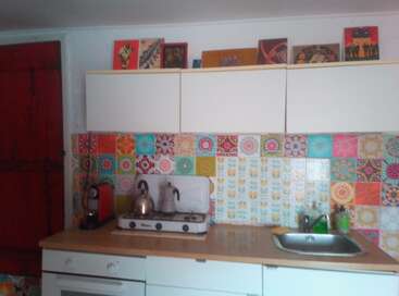 This kitchen features colorful, patterned tile backsplash, a countertop stove with kettles, a coffee maker, white cabinets, a sink, and decorative art on top cabinets.