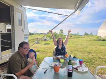 A family enjoys an outdoor meal beside their RV on a sunny day. A man holds a baby, while a woman raises her arms, celebrating.