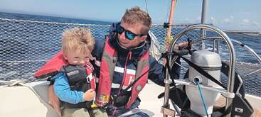 A man and a young boy, both wearing life jackets, are sailing on a boat. The man is steering, and they are smiling, enjoying sunny weather.