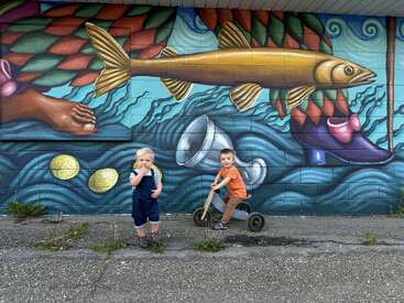 Two young children pose in front of a colorful mural featuring a large yellow fish, floating coins, shoes, a cup, and textured oceanic patterns.