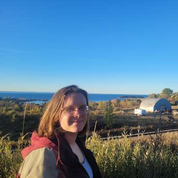 A person with shoulder-length hair and glasses stands outdoors with lush greenery, a blue sky, distant water, trees, and a white building in the background.