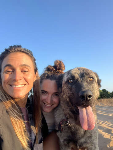 Two women and a large, happy dog are enjoying a sunny day outdoors, smiling at the camera with clear blue skies in the background. Pure joy!