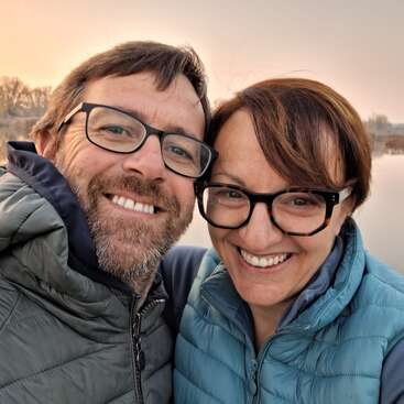 Ein lächelnder Mann und eine lächelnde Frau mit Brille und Winterjacken machen draußen ein Selfie aus nächster Nähe. Im Hintergrund sieht man einen ruhigen See und sanftes, warmes Abendlicht.