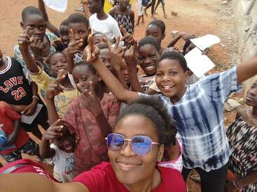 A group of joyful children gather closely for a selfie with a smiling woman. Everyone looks excited, happily posing with playful gestures and cheerful expressions outdoors.