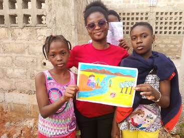 Three girls pose outside with a colorful landscape drawing featuring a river, trees, and mountains. Two girls hold the artwork proudly, while another stands behind them.