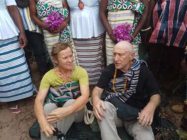 A group photo featuring two seated people, a woman and a man, surrounded by others standing, all wearing colorful, traditional patterned clothing and holding purple flowers.
