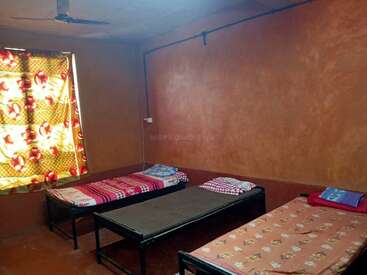 Esta imagem mostra um quarto simples com três camas de solteiro de metal, roupas de cama coloridas, uma cortina floral, paredes marrons, um ventilador de teto e decoração minimalista.