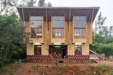 Uma casa moderna de dois andares com janelas grandes, paredes marrons e um telhado inclinado, cercada por vegetação e árvores; espaço inferior aberto com móveis visíveis.