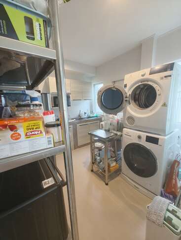 L'image montre une buanderie avec un lave-linge et un sèche-linge empilés, des portes ouvertes, une étagère métallique avec des fournitures, et un petit coin cuisine organisé.