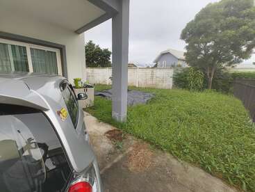 Une voiture argentée est garée à côté d'une maison. Le jardin est envahi par l'herbe et un grand arbre. Une bâche grise recouvre une partie de la cour.