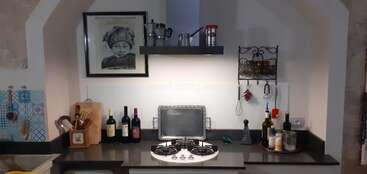Essa cozinha aconchegante tem fogão a gás, garrafas de vinho, utensílios de cozinha, temperos, garrafas de óleo, uma prateleira de parede e um retrato emoldurado acima da bancada.