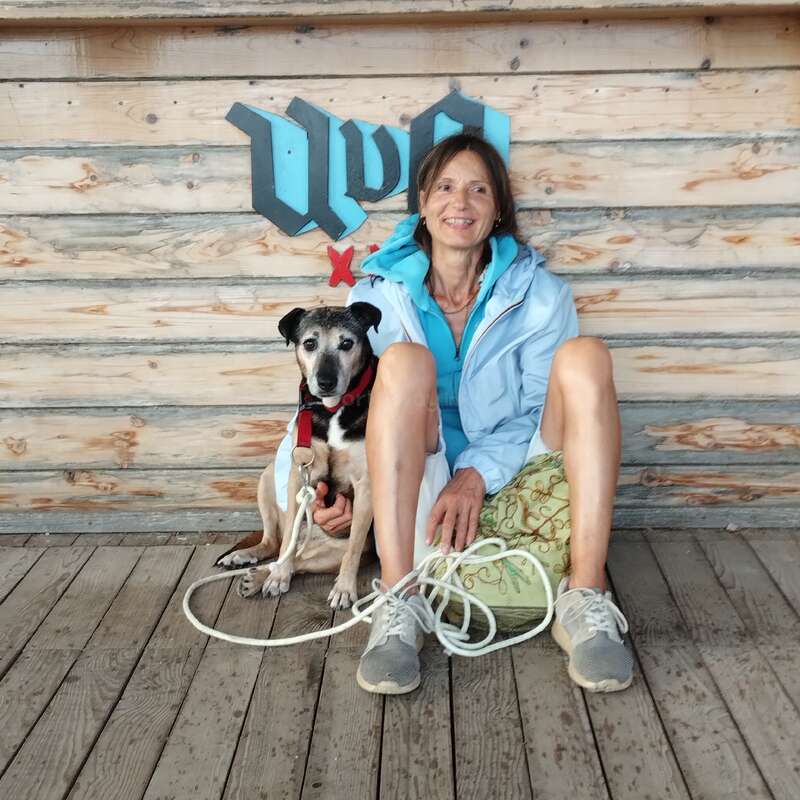 Uma mulher sorridente com uma jaqueta azul clara está sentada no chão de madeira ao lado do seu cachorro. Eles estão em frente a uma parede de madeira com grafites azuis.