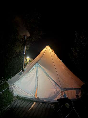 Une grande tente en toile éclairée se dresse sous un ciel nocturne sombre. De la fumée s'échappe d'une cheminée, laissant deviner la chaleur qui règne à l'intérieur. Des arbres et du matériel de camping l'entourent dans une atmosphère paisible.