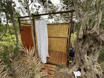 Cette image montre une douche extérieure rustique avec des parois en bois, un fond en bambou, un rideau blanc et un sol en caillebotis, le tout entouré d'arbres et de verdure.