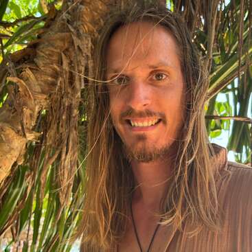A smiling man with long blond hair and a beard stands outdoors, surrounded by tropical greenery and palm trees, wearing a brown shirt and a necklace.