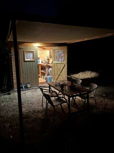Un cobertizo de madera con una cálida iluminación y las puertas abiertas por la noche, que deja entrever un interior abarrotado. En el exterior, hay una mesa con cuatro sillas bajo un toldo.