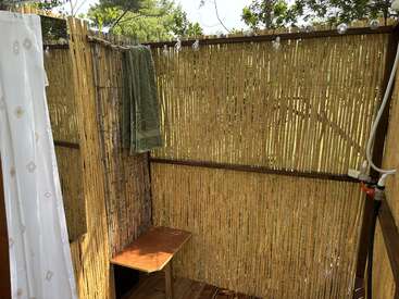 Se trata de una ducha al aire libre con paredes de bambú, un banco de madera, una toalla verde, guirnaldas de luces y una cortina blanca, rodeada de vegetación y luz natural.