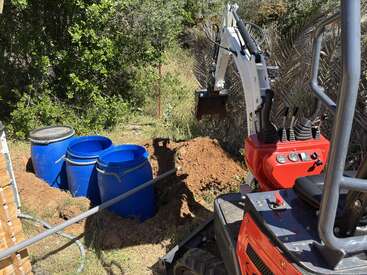 Una pequeña excavadora está excavando junto a unos bidones azules, quizá para una obra o un proyecto de fontanería, rodeada de árboles y vegetación en una zona al aire libre muy soleada.