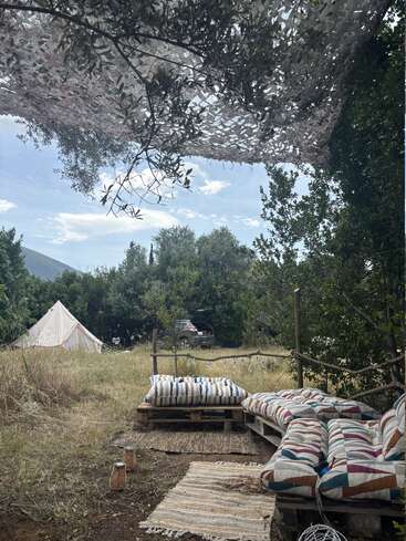 Una acogedora zona de descanso al aire libre, con cojines de colores sobre palés de madera y alfombras en el suelo, a la sombra de una malla, desde la que se divisa una tienda de campaña y un coche entre la exuberante vegetación.