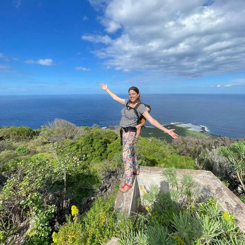 A imagem mostra uma mulher em pé em um parapeito de concreto, com os braços estendidos, com vista para um vasto corpo de água e uma vegetação exuberante sob um céu azul com nuvens brancas.