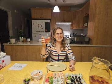 Une femme souriante est assise à une table de cuisine avec des sushis, des baguettes et une boisson. Elle lève son verre pour porter un toast, profitant d'un dîner confortable à la maison.