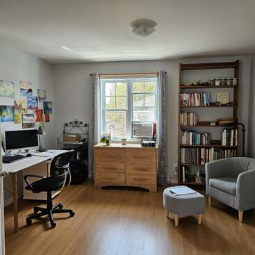 Un bureau confortable comprend un bureau avec un ordinateur, une chaise pivotante, une bibliothèque remplie de livres, une fenêtre avec des rideaux, un fauteuil confortable, un pouf et des œuvres d'art.