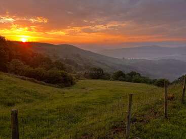 L'image représente un coucher de soleil serein sur des collines ondulantes, avec une clôture métallique au premier plan et un paysage verdoyant avec des arbres et des collines à l'arrière-plan.