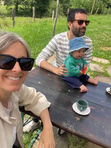 L'image représente une famille de trois personnes assises à une table de pique-nique dans un champ herbeux, avec un homme, une femme et un bébé profitant ensemble d'une journée ensoleillée.