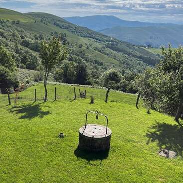 L'image représente un champ herbeux serein avec un puits, des arbres et une clôture, avec en toile de fond des collines et des montagnes ondulantes sous un ciel nuageux.