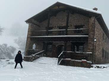 Une personne marche dans la neige devant une maison en pierre de deux étages avec un balcon et des escaliers, par une journée enneigée.