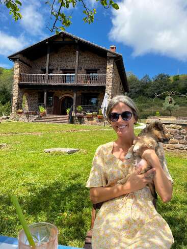 Das Bild zeigt eine Frau, die in einem Stuhl sitzt und einen kleinen Hund vor einem rustikalen Steinhaus an einem sonnigen Tag mit einem ruhigen, grasbewachsenen Hof und blauem Himmel wiegt.
