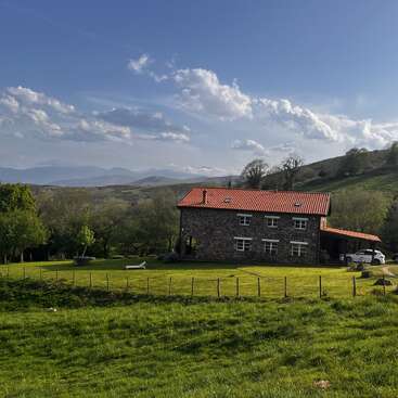 A rustic stone house with a red roof sits in a lush green meadow, surrounded by trees, hills, and distant mountains under a vibrant blue sky.
