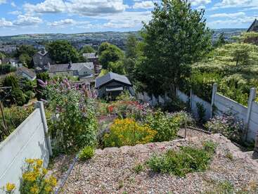 Dieses Bild zeigt einen bunten, blühenden Garten auf einem Hügel, umgeben von weißen Zäunen, mit Blick auf Häuser, Bäume und eine malerische, hügelige Landschaft unter einem blauen Himmel.