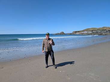 Ein Mann mit einem karierten Hemd und einer Mütze steht an einem Sandstrand und hält ein Getränk in der Hand. Das Meer und die felsige Küste liegen unter einem klaren blauen Himmel.