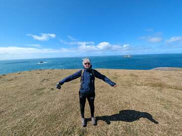 Eine Person steht mit ausgestreckten Armen auf einer grasbewachsenen Klippe und blickt auf einen weiten blauen Ozean unter einem klaren, sonnigen Himmel mit vereinzelten Wolken.