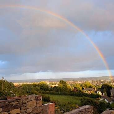 Ein leuchtender Regenbogen spannt sich über einen wolkenverhangenen Himmel über einer friedlichen Stadt. Üppig grüne Bäume und Häuser füllen die ruhige Landschaft unter dem farbenfrohen Spektakel.