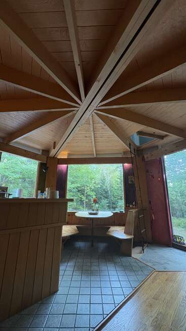 O interior aconchegante da cabana de madeira tem um teto radial único, janelas grandes com vista para a vegetação exuberante, uma mesa redonda com bancos e um ambiente simples e tranquilo.