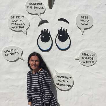 La imagen muestra a una mujer de pie frente a una pared blanca adornada con ojos de dibujos animados y bocadillos con texto en español, probablemente para promocionar un producto o servicio.