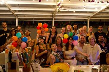 Um grupo alegre de pessoas se reúne sob luzes de cordas, sorrindo e rindo. Alguns seguram balões coloridos e flores, criando uma atmosfera festiva, calorosa e comemorativa.