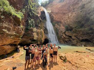 Um grupo de amigos posa alegremente em frente a uma cachoeira cênica cercada por penhascos rochosos, água limpa e vegetação exuberante sob a luz do dia.