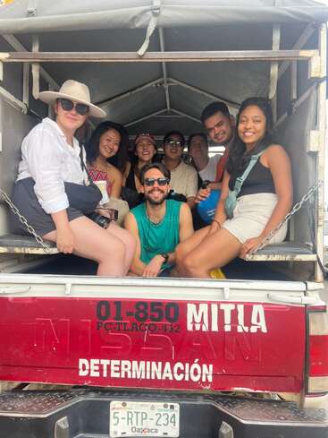 Um grupo de nove amigos se senta e sorri dentro de um caminhão, aproveitando um passeio juntos. O ambiente é descontraído e alegre, com roupas e chapéus de verão.